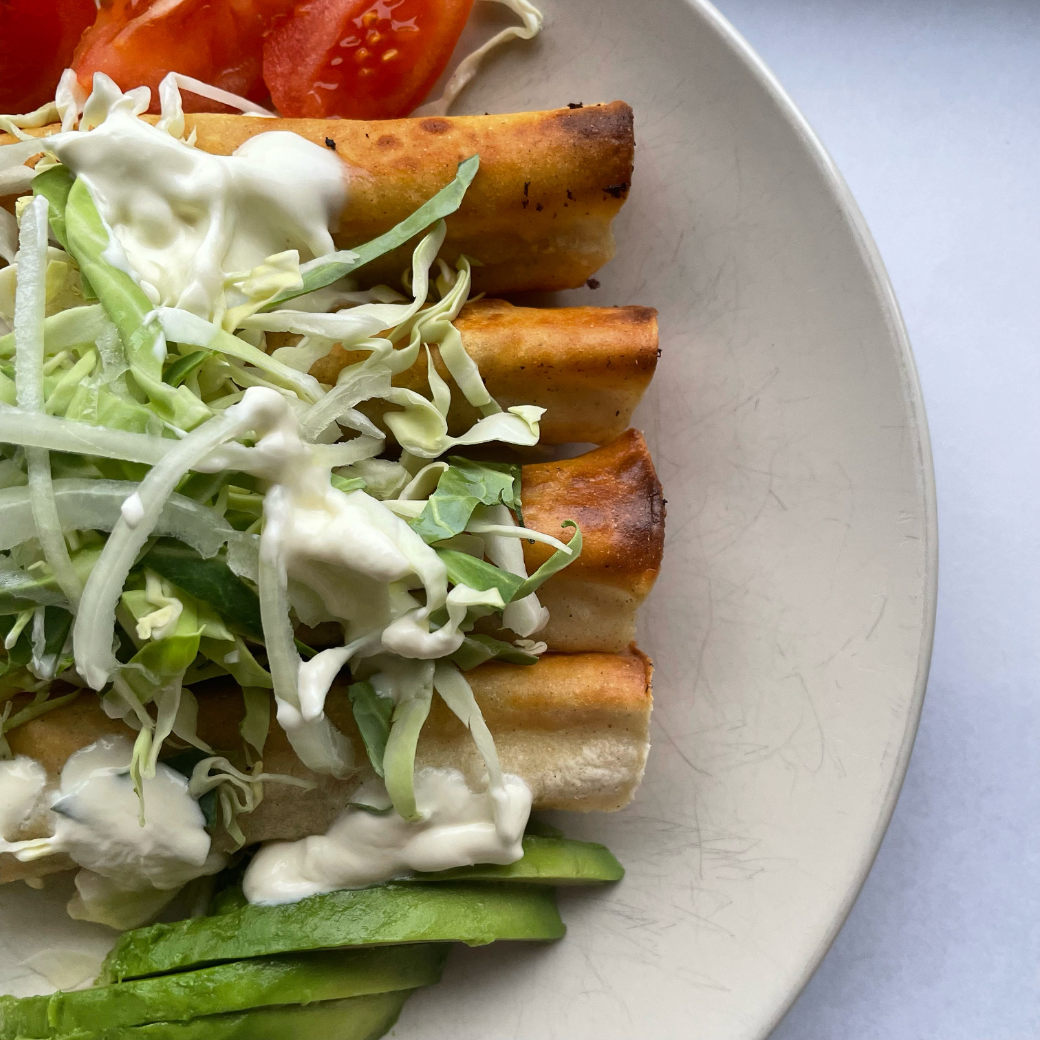 Rolled tacos surrounded by sliced avocado, tomatoes, cabbage, onion, and sour cream